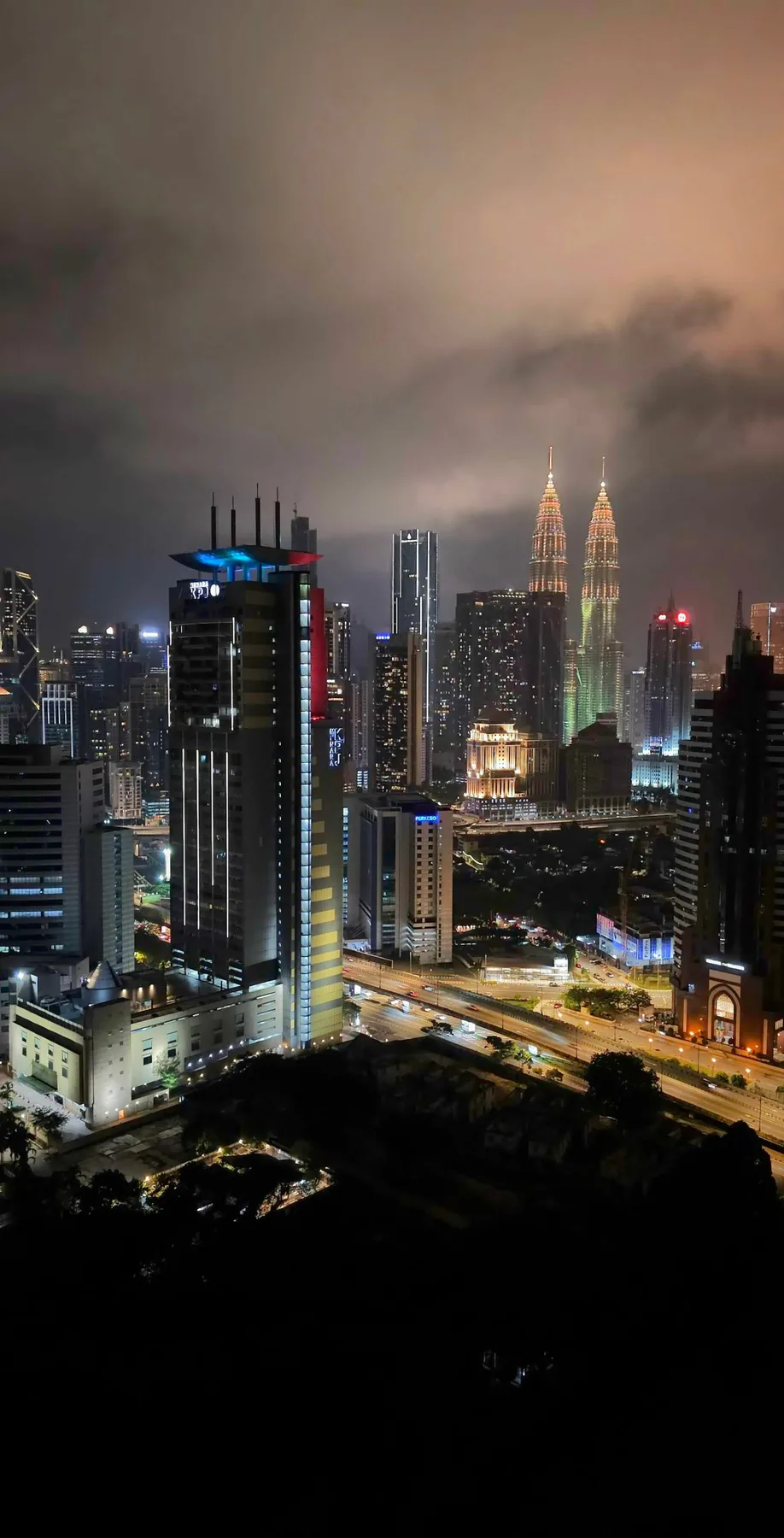 Geceleri aydınlatılan Kuala Lumpur binaları ve uzaktaki ikiz kuleler.